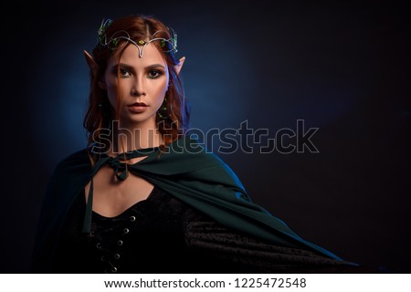 Crop of young and beautiful woman in green cloak posing at camera. Charming queen of elves in silver tiara and red hair gesturing by her left hand. Isolated of girl on dark blue studio background.