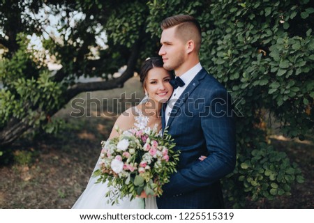 Beautiful newlyweds cuddle in a green garden with trees. Young bride and cute bride with a bouquet are standing in the park. Wedding photography. Stylish portrait.