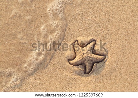 Starfish drawn on the beach sand being washed away by a wave. Foaming sea wave coming to wash a picture on wet yellow beach sand. Holiday and vocation message. View from above. Copy space.