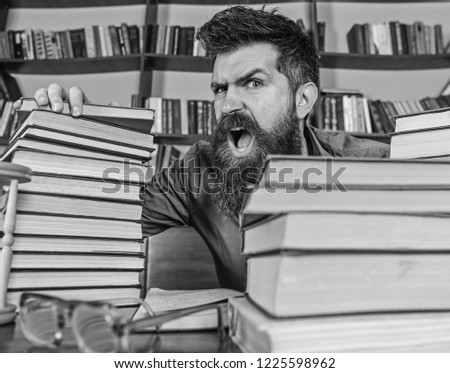 Teacher or student with beard sits at table with glasses, defocused. Man on shouting face between piles of books, while studying in library, bookshelves on background. Mad scientist concept.