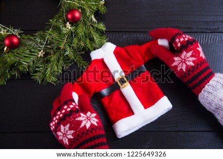 Hands in red winter gloves holding toy Santa costume on black wooden background. New Year decoration with fir wreath and Christmas balls.