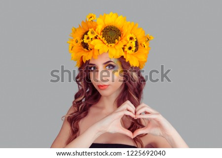 Pretty woman with floral headband making a heart gesture with her fingers in front of her chest showing her love, affection with a happy tender smile. Fashion girl crown from sunflower on head on gray