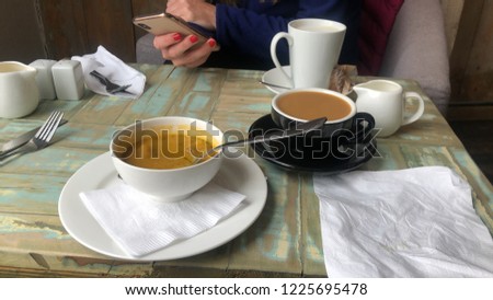 Vegetable Soup and fresh coffee and soya milk on a table with female with red nail polish holding and typing on a smart phone