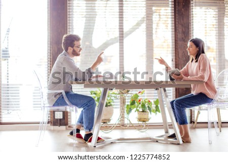 Young handsome businessman talking with his wife, while drinking coffee, sitting opposite at the same table in light airy interior with big windows. Copy space