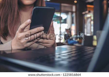 Closeup image of a woman holding , using and looking at smart phone with laptop on table in cafe