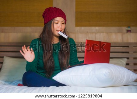 lifestyle home portrait of young beautiful and happy Asian Japanese woman in winter hat sitting relaxed on bed holding credit card using laptop computer for internet banking and online shopping 