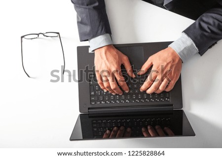 Man with laptop typing on the keyboard. On white table. The man wears a suit and a shirt. Eyeglasses resting on the table.