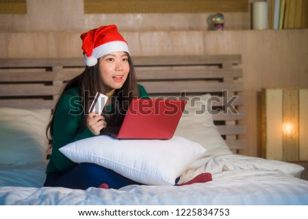 young happy and beautiful Asian Korean girl in Santa Christmas hat using credit card and laptop computer for online shopping x-mas presents and gifts sitting joyful and excited on bed 