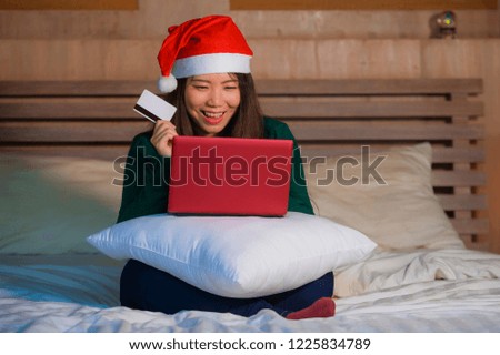young happy and beautiful Asian Chinese girl in Santa Christmas hat using credit card and laptop computer for online shopping x-mas presents and gifts sitting joyful and excited on bed 