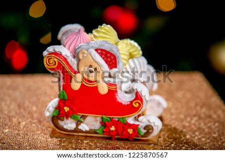 Gingerbread sleigh with teddy bear in front of defocused lights of Christmas decorated fir tree. Holiday sweets. New Year and Christmas theme. Festive mood.