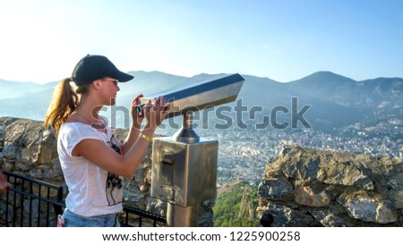 Young Girl - traveler, looking through Binocular ,  panoramic viewing of Alanya city and beaches from the viewpoint of the mountain: "Alanya Kalesi". Toned picture, Copy space for your design Royalty-Free Stock Photo #1225900258