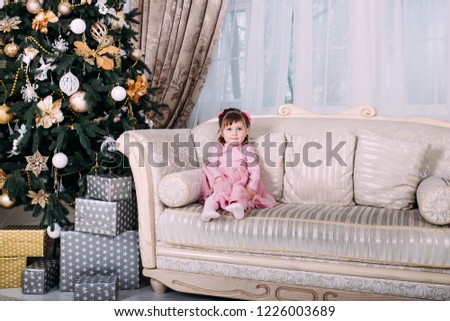 Little girl sitting on sofa near Christmas tree indoors. Merry Christmas and Happy Holidays!