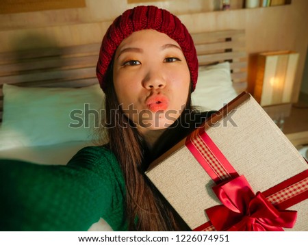 young beautiful and happy Asian American girl taking selfie picture with mobile phone holding Christmas gift box smiling cheerful and excited in winter beamy at home celebrating xmas holidays