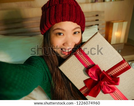 young beautiful and happy Asian American woman taking selfie picture with mobile phone holding Christmas gift box smiling cheerful and excited in winter beamy at home celebrating xmas holidays