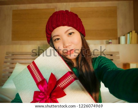 young beautiful and happy Asian Korean woman taking selfie picture with mobile phone holding Christmas gift box smiling cheerful and excited in winter beamy at home celebrating xmas holidays