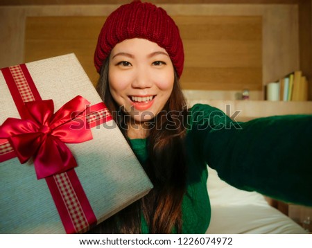 young beautiful and happy Asian Chinese woman taking selfie picture with mobile phone holding Christmas gift box smiling cheerful and excited in winter beamy at home celebrating xmas holidays