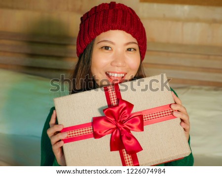 young happy and beautiful Asian Chinese girl in winter hat holding Christmas present box with ribbon smiling excited and cheerful sitting at home bed celebrating xmas receiving gift