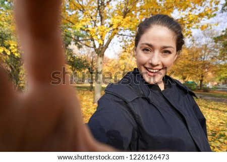 happy young woman in her 20s taking selfie photo posing in park with autumn colors - wide angle shot with invisible smartphone