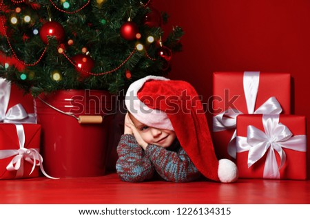 Happy xmas and New Year. Portrait of child in Santa red hat waiting for Christmas gifts. Little baby boy lies under the Christmas tree with presents and is waiting for a miracle