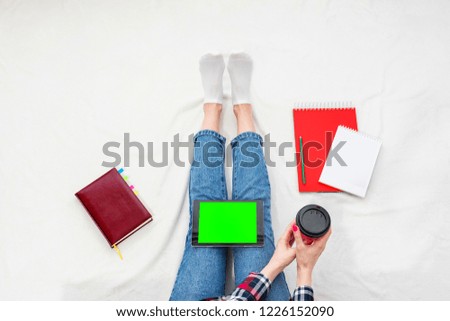 Girl in blue jeans and plaid shirt, woman holds in her hands coffee from disposable cup, tablet pc, women's feet, lying on white crumpled blanket. Women's hands. Background with copy space