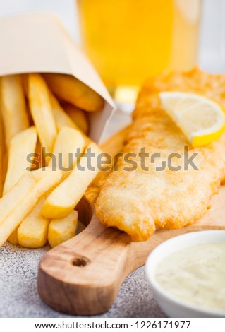 Traditional British Fish and Chips with tartar sauce abd glass of craft lager beer on chopping board on white stone background.