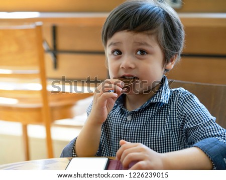 Portrait of happy boy eating chocolated in the coffee shop while playing game on mobile phone,Kid enjoying time watching cartoon and eating sweet snack in the cafe, Positive kid concept