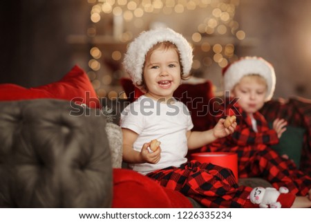 Two small cute children in Santa's hats are sitting on a soft home sofa and having fun. Home Family Christmas Holiday