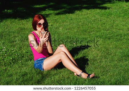 A beautiful girl with red long hair is sitting on the grass in the park, she has a phone in her hand. Emotional photo