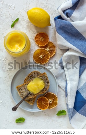 Slice of wholegrain bread with lemon custard, lemon and a glass jar with lemon custard on a textured white background and gray linen cloth. Top view.