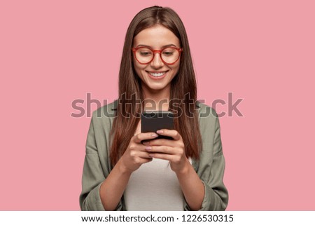 Satisfied lovely woman holds modern cell phone, texts with friend online in chat, dressed in stylish clothes, poses against pink wall, connected to high speed internet. Shot of teenager with device Royalty-Free Stock Photo #1226530315