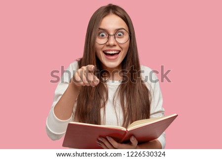 Lovely glad surprised girl smiles broadly and points directly into camera, wears transparent glasses, holds red book, notices something in distance, isolated over pink background. Wow, look there