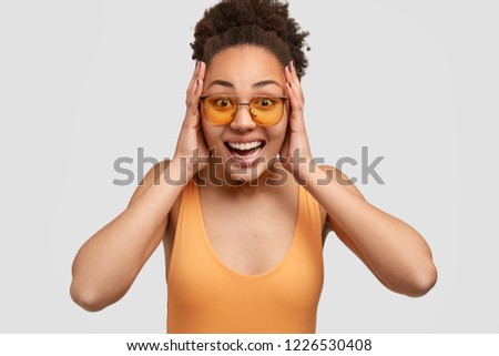 Thrilled overjoyed dark skinned girl keeps both palms on head, feels excited while talks with friend about something awesome, dressed in casual clothing, poses over white background. Emotions concept