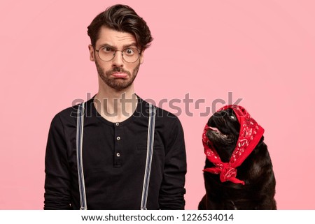 Portrait of displeased frustrated man with negative sullen facial expression, purses lower lip in bewilderment, discontent with something, black dog wears bandana on head, looks at its owner