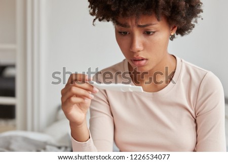 Embrarrassed woman with Afro hairstyle faces serious problem, dissatisfied with pregnancy test, dressed in pyjamas, has displeased expression, models indoor, thinks about abortion. How it possible?