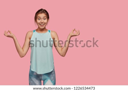 Horizontal portrait of amazed female meditates indoor, keeps both hands in okay gesture, tries to relax, prepares for sport competitions, dressed in sportsclothes, isolated on pink background