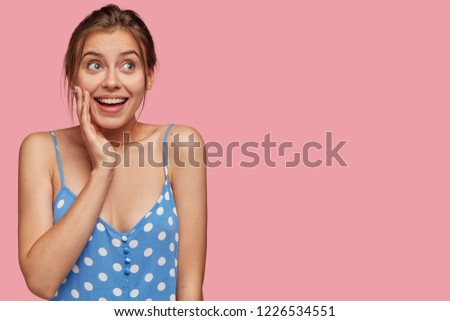 Picture of beautiful Caucasian woman looks joyfully upwards, keeps hand on cheek, being glad, dreams about something pleasant, dressed in blue polka dot dress, stands against pink studio wall