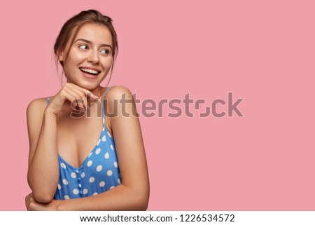 Indoor shot of happy young Caucasian woman keeps hand under chin, looks joyfully at camera, has healthy skin, shows natural beauty, being in good mood after date, isolated over pink background