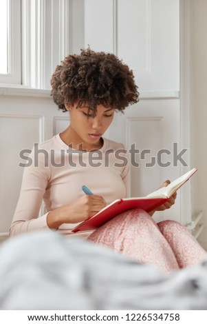 Photo of beautiful calm copywriter writes down information in notebook, wears pyjamas, works from home before sleep, focused on work. Creative journalist or writer records something in organizer