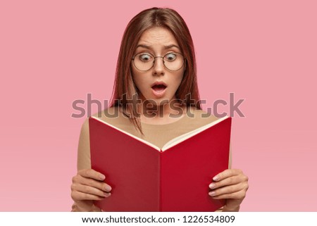 Headshot of surprised woman with terrified expression, stares at book, reads shocking information while prepares for examination, keeps jaw dropped, isolated over pink background. Reaction concept