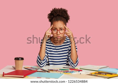 Dissatisfied dark skinned woman keeps index fingers on temples, gathers with thoughts, dressed in striped clothes, surrounded with crayons and blank sheet of papers. People and work concept.