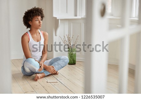 Photo of concentrated black woman sits crossed legs on wooden floor, has thoughtful expression, meditates indoor, modern cell phone near, listens calm pleasant music. People and wellbeing concept