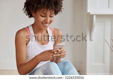 Positive African female enjoys leisure time, uses modern technology, addicted to social networks, wears white vest and leggings, chats with followers, connected to wireless internet, texts message
