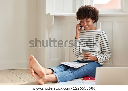 Glad dark skinned smiling woman with Afro hairstyle, discusses brokerage services via smart phone, accounts budget, has telephone conversation with business associate, holds paper cup of coffee
