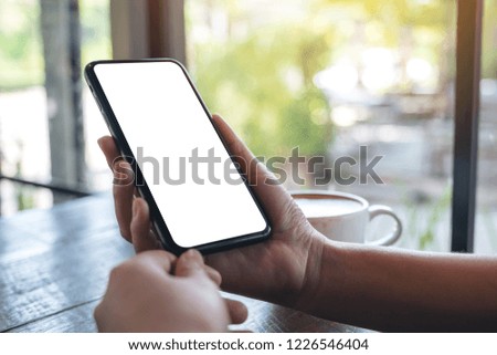 Mockup image of a woman's hands holding black mobile phone with blank desktop screen with coffee cup on wooden table in cafe