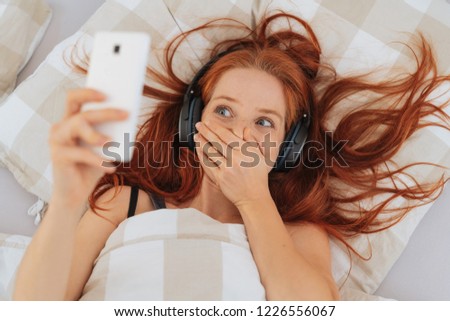 Embarrassed young redhead woman covering her mouth as she looks at something on her mobile phone with lying in bed listening to music