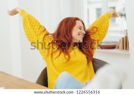 Happy pretty young woman stretching her arms as she leans back relaxing in her chair with a beaming smile Royalty-Free Stock Photo #1226556316