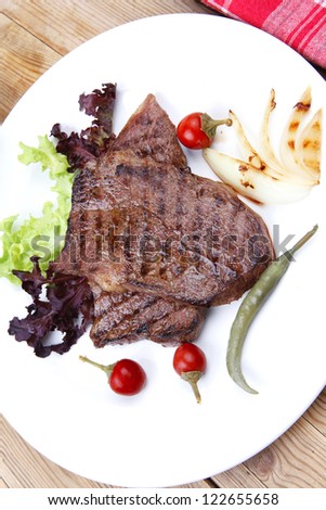 meat food : two grilled steak on green lettuce salad , with roast onion and red hot chili peppers , on dish over wooden table