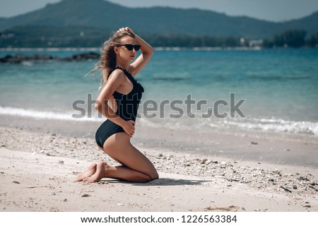 Young fit woman in a black swimsuit and black sunglasses sitting on the wet sand beach . Karon beach. Phuket.