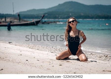 Young fit woman in a black swimsuit and black sunglasses sitting on the wet sand beach with out of focus boat in background. Karon beach. Phuket.
