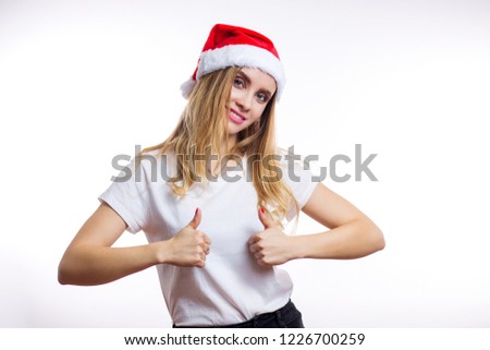 Happy charming blonde girl in red Santa hat and t-shirt shows sign thumb up with two hands and smiles at camera on white background. Christmas, new year, Expressive facial expressions emotions concept